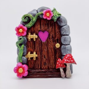 C'est un magnet porte de fée, en forme de porte en bois avec un coeur en son centre, avec une arche en pierre, deux champignons rouge se trouvent en bas à droite, et sur la moitié gauche de l'arche se trouve une liane verte avec 3 fleurs roses