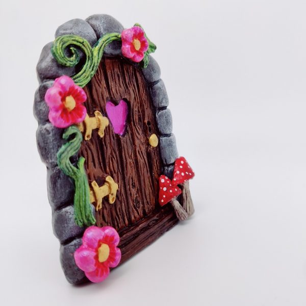 C'est un magnet porte de fée, en forme de porte en bois avec un coeur en son centre, avec une arche en pierre, deux champignons rouge se trouvent en bas à droite, et sur la moitié gauche de l'arche se trouve une liane verte avec 3 fleurs roses