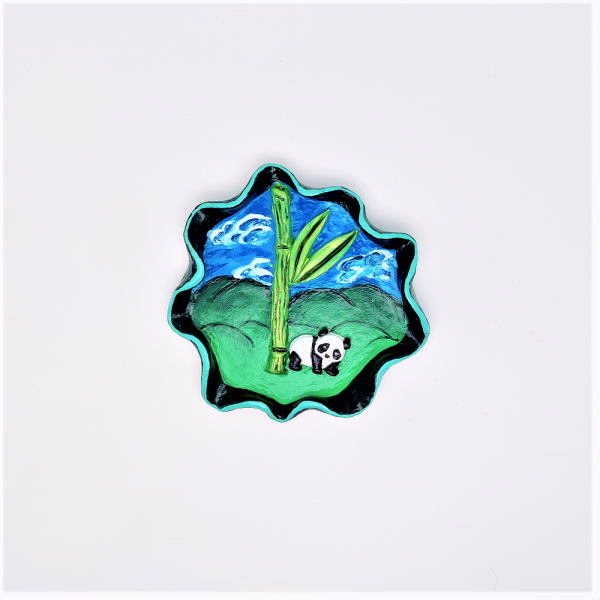 Coupelle à bijoux en argile peint. Rebord ondulé avec un liseré vert clair. En fond des collines et un ciel bleu. En relief un bambou et un panda.