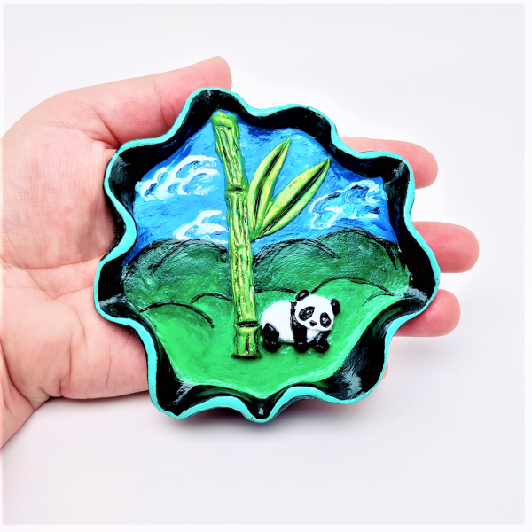 Coupelle à bijoux en argile peint. Rebord ondulé avec un liseré vert clair. En fond des collines et un ciel bleu. En relief un bambou et un panda.