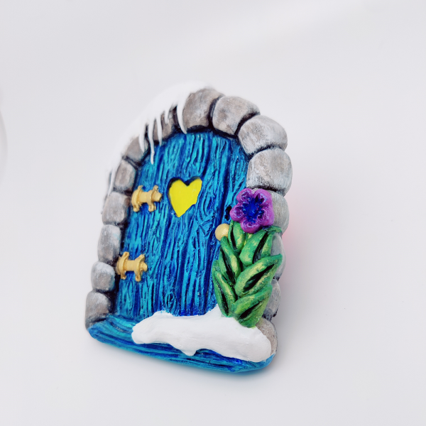 Magnet porte de fée enneigée. Arche de pierre entourant une porte en bois bleu glaciale avec de la neige tombante en haut à gauche. De la neige accumulé en bas à droite et une fleur bleue violette avec des feuilles vertes irrisées
