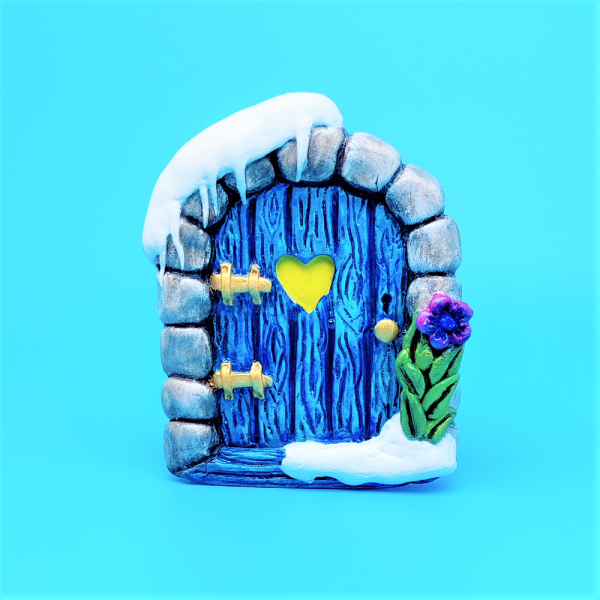 Magnet porte de fée enneigée. Arche de pierre entourant une porte en bois bleu glaciale avec de la neige tombante en haut à gauche. De la neige accumulé en bas à droite et une fleur bleue violette avec des feuilles vertes irrisées